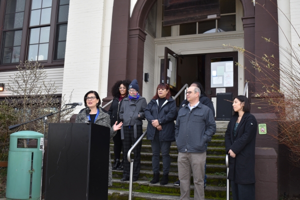 Councilmember Tammy Morales stands at a podium in front of the Beacon Hill School, surrounded by supporters.