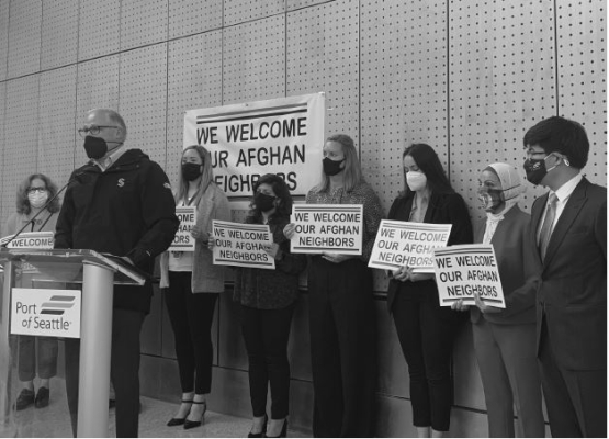 Governor Inslee, dressed in dark outerwear jacket and face mask, stands at podium in front of seven people, most holding signs that say, "We Welcome Our Afghan Neighbors."