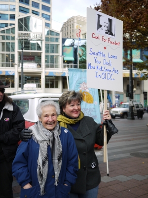 Two women stand together on city street, one holding a sign reading, "Dorli for President. Seattle Loves You Dorli, Now Kick Some Ass in Ol' DC."