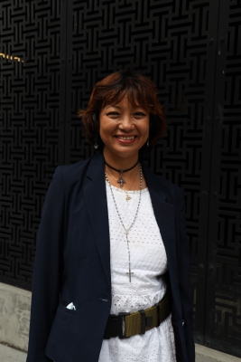 Full-length photo of smiling middle-aged Asian woman with chin-length light brown hair, dressed in suit jacket over white dress and two necklaces with crosses