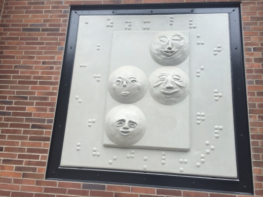 Wall hanging showing Braille letters behind inset with four clay faces