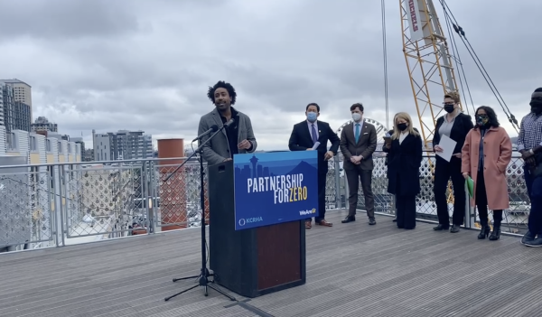Young Black man stands at podium on a rooftop, with row of other people standing to the side, with cloudy sky and crane in background.
