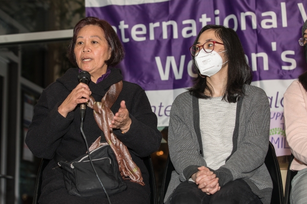 Two Filipina women, older one holding mic, sitting in front of purple International Women's Day sign