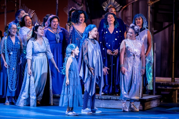 Women and girls of all ages, dressed in long blue dresses, stand on stage singing.