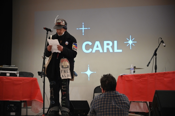 Carl Nakajima, dressed in cap and jacket decorated with badges, stands at podium holding sheet of paper, in front of screen reading "Carl" surrounded by stars