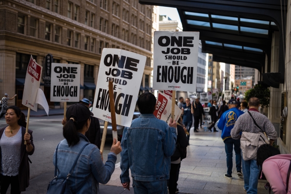 People walk along city sidewalk holding signs that read, "One Job Should Be Enough."
