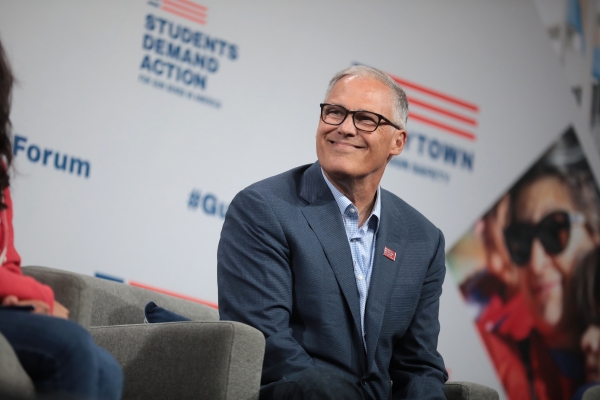 Governor Jay Inslee, dressed in checked shirt and wool suit jacket, seated in a chair at a conference event, smiling