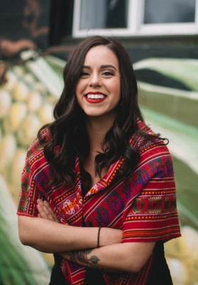 Headshot of young, dark-haired woman in red serape-style dress standing in outdoor environment
