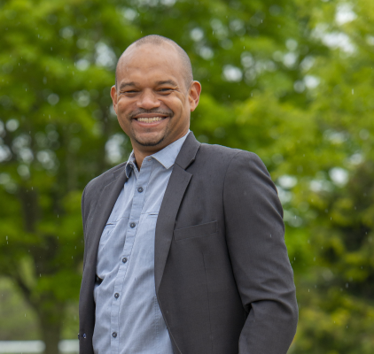 Photograph of smiling Black man standing outdoors in shirt and jacket 