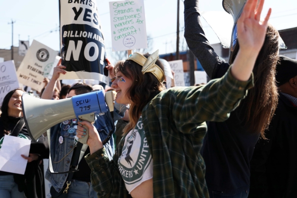 Young white woman wearing flannel shirt over cut-off Workers United T-shirt holds megaphone in one hand and gestures with the other, surrounded by a rally crowd.