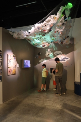 Two children and an adult man stand in a gallery space, walls covered with cutouts of sea animals and an installation hanging from ceiling.