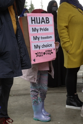 Child in purple rainboots and colorful leggings holds pink sign in front of top half of body, reading "Hijab, My Pride, My Freedom, My Choice"