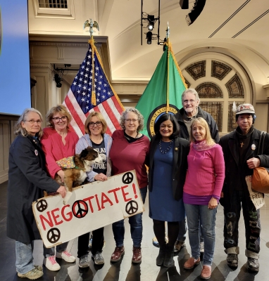 Group of people, including Medea Benjamin, Pramila Jayapal, Carl Nakajima, and three women holding a dog and a sign reading "Negotiate!" stand in a lobby in front of American flag and Washington state flag.
