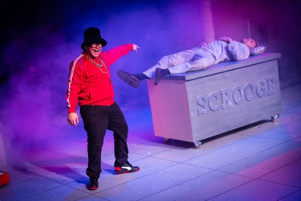 Goofy figure in red shirt, hip-hop style gold chain necklace, and black top hat smiles and points to a box showing word "Scrooge" on the side, with man in pajamas sleeping on top of it.