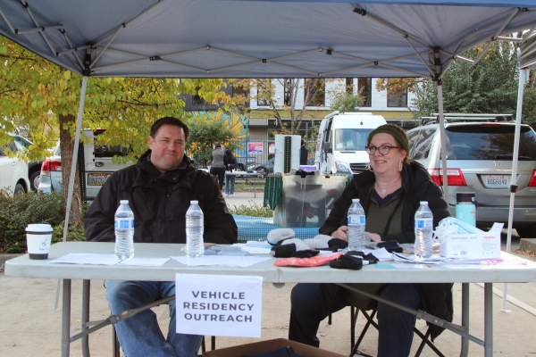 Two people, middle-aged white man and woman, sit side by side at table laden with water bottles and socks, with sign hanging from edge reading, "Vehicle Residency Outreach."