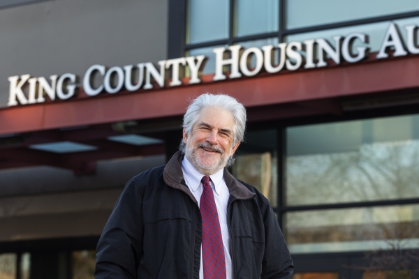 Man with white hair, beard, and mustache, dressed in shirt and tie under barn jacket, stands in front of building with County Housing sign