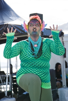 Brown-skinned person with dragon makeup and green-mesh top and pants strikes a monster pose with hands in air.