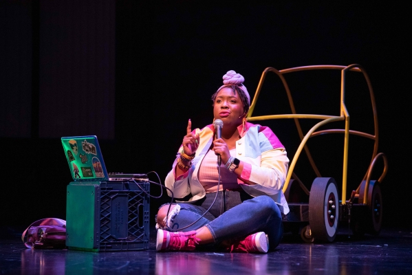 The actor playing Lydia sits on stage in front of a toy-sized car holding a microphone connected to an amp.