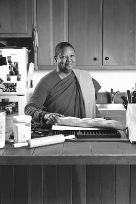 The Venerable Pannavati, the world's only black Buddhist nun, is in a kitchen. She has one hand placed on a loaf of bread. In the foreground is jar and rolling pin, and a fridge in the background.