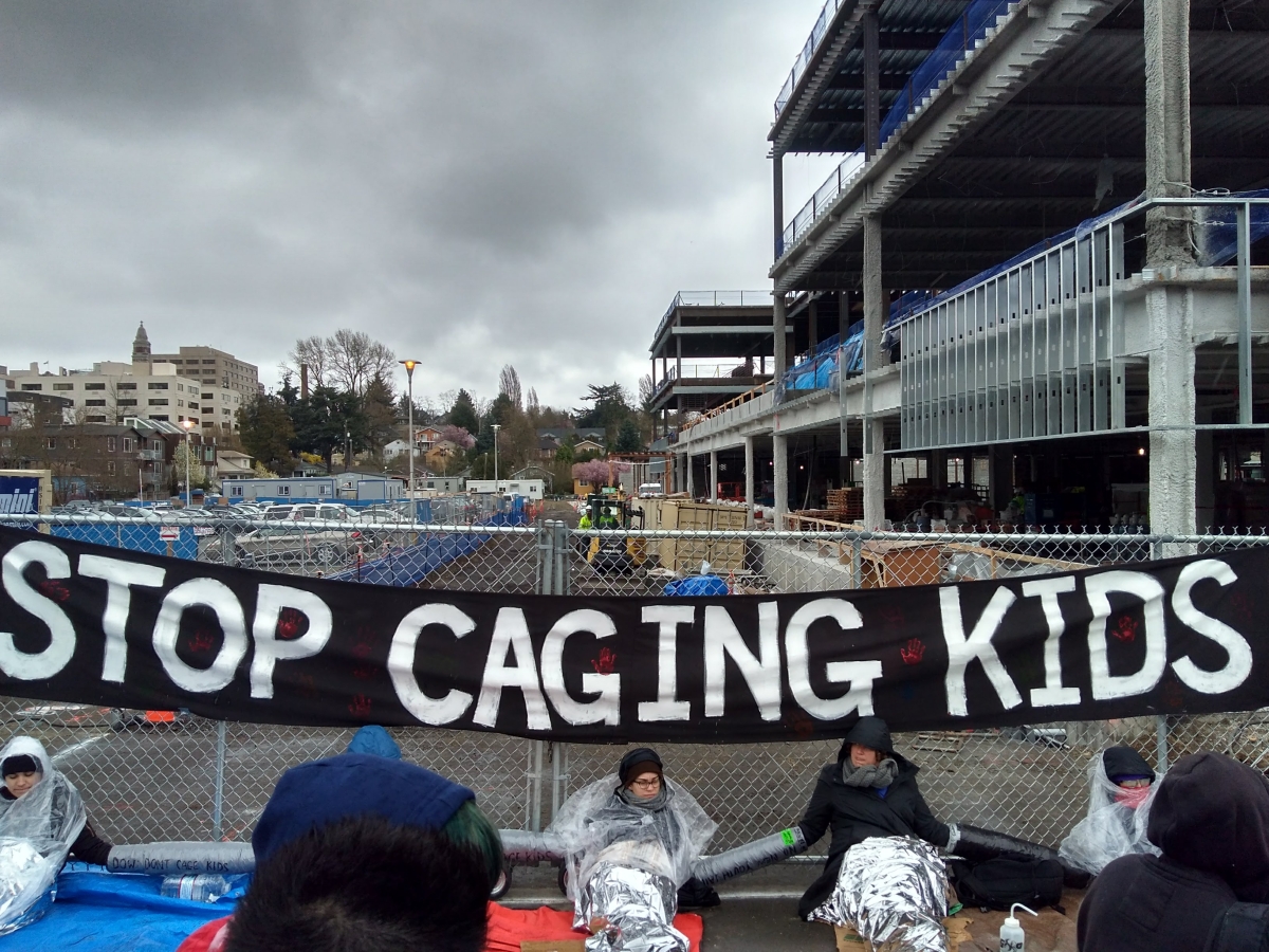 A photo of people protesting against the new King County youth jail while it was being constructed. On the fence behind the people is a banner labeled "Stop Caging Kids"
