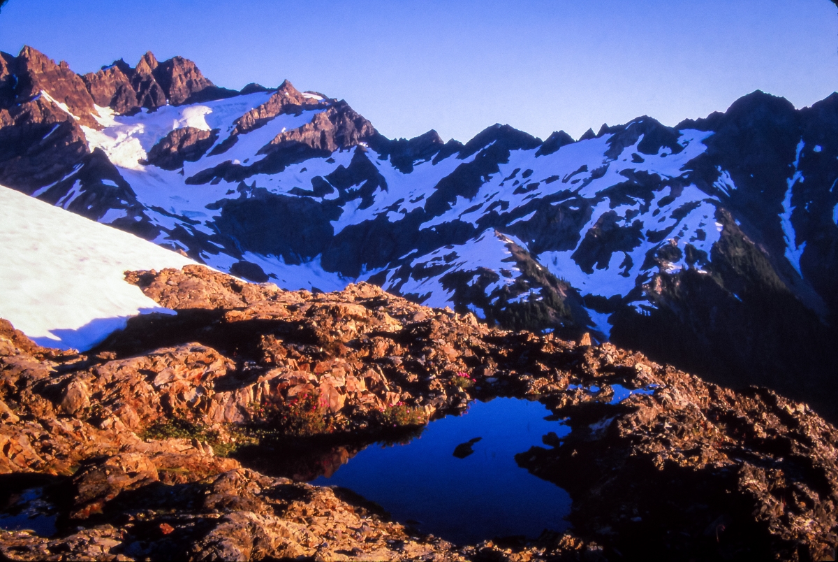 Photograph of mountains against sunny sky, snow-capped in background and circling small lake in foreground