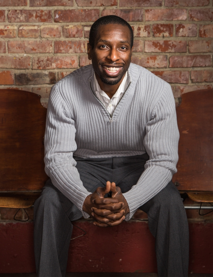 Young Black man in light-colored crewneck sweater and dress pants, sitting in front of brick wall