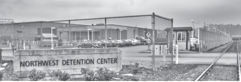 Black and white photo with Northwest Detention Center sign in front of fence and parking lot
