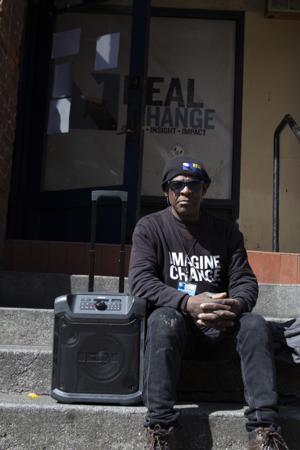 Black man sits on steps next to a portable speaker, wearing cap and sweatshirt that reads, "Imagine Change."