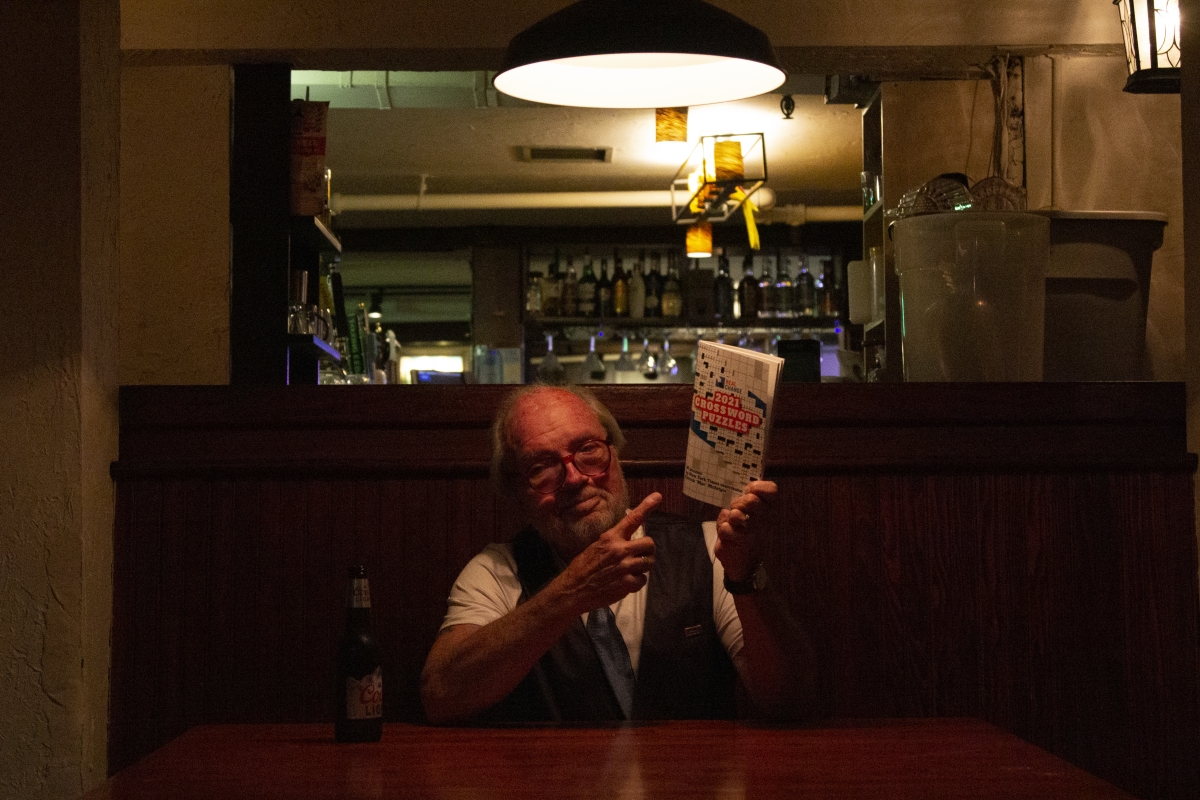 A photo of an older man holding up a copy of the Real Change crossword book while drinking a Coors Light in a wood-paneled bar