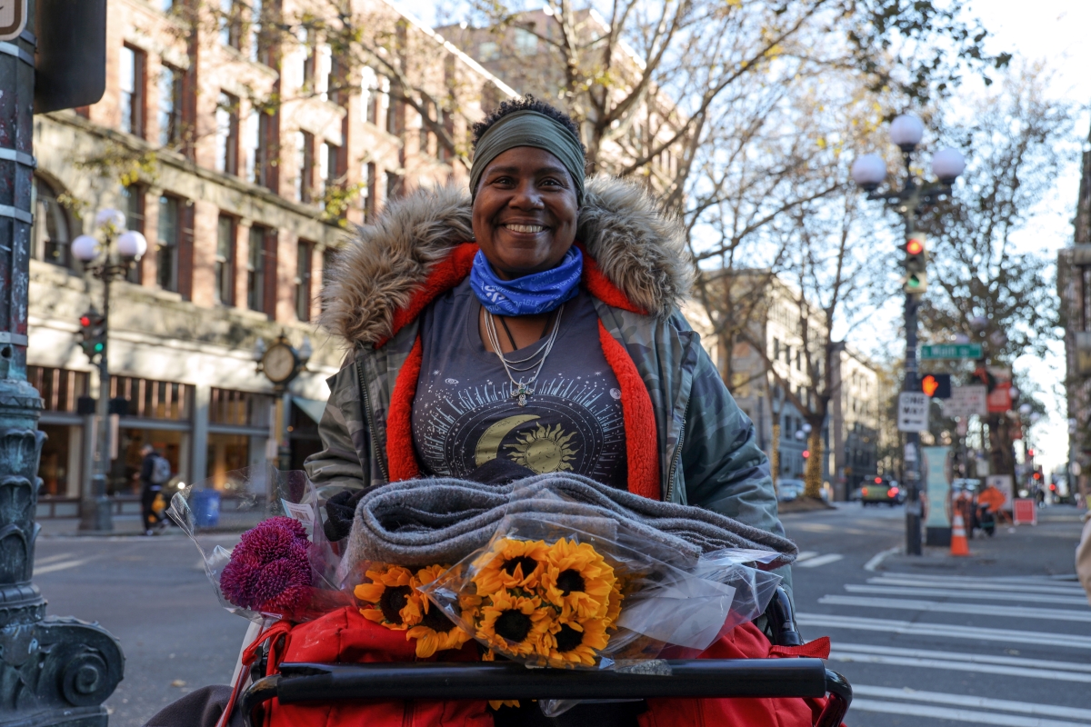 Smiling youngish Black woman in coat with fur-looking collar, gray headband, and colorful T-shirt holds folded blankets and flowers.