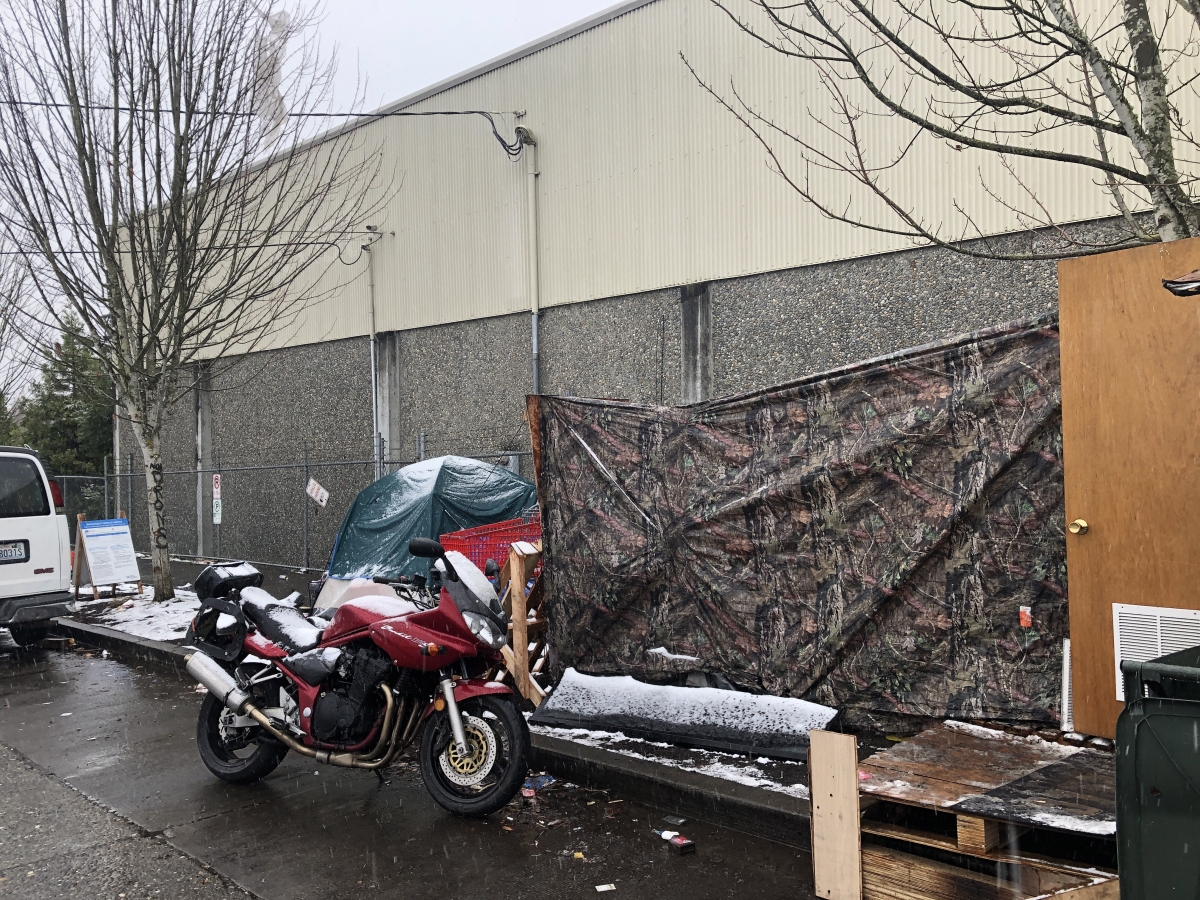 Photograph of side of a gray building in parking lot on wet winter day, with tent tarp strung up behind motorcycle in foreground