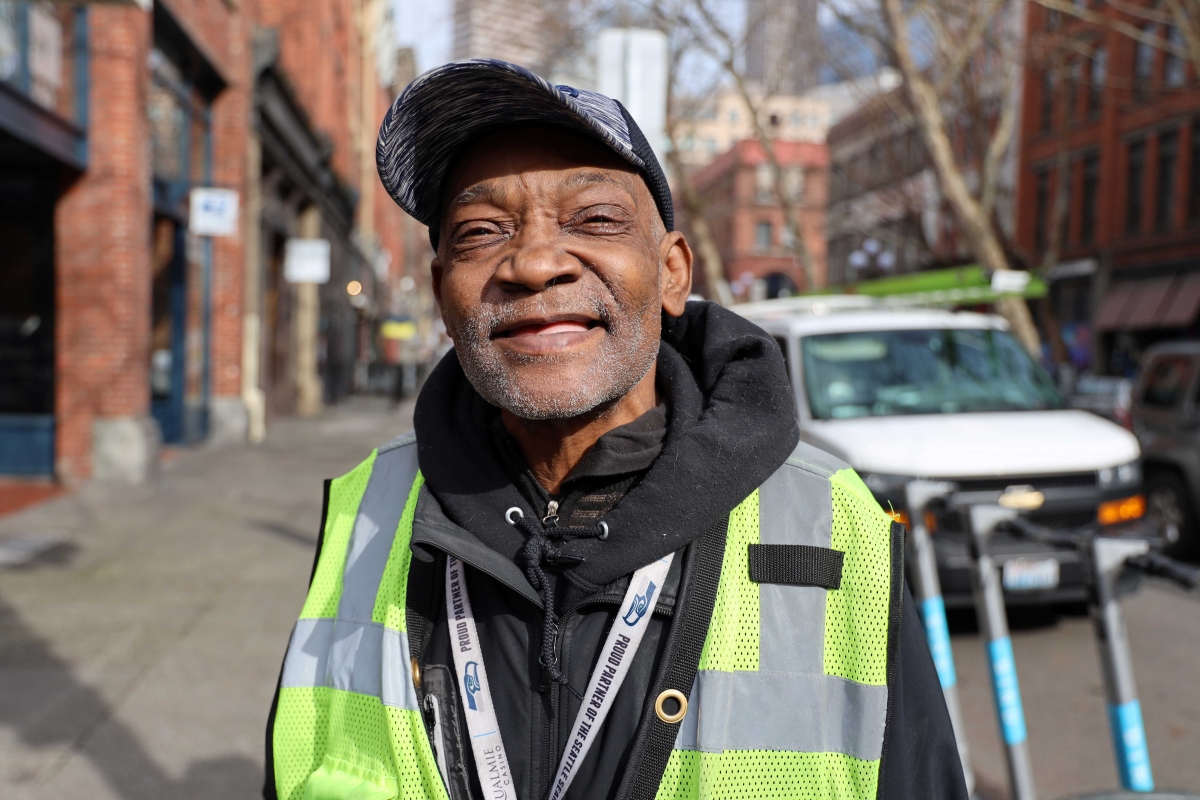 A portrait of vendor Canada Jones, an older Black man wearing a hoodie with his bright yellow reflective Real Change vest over it, in front of the Real Change offices.