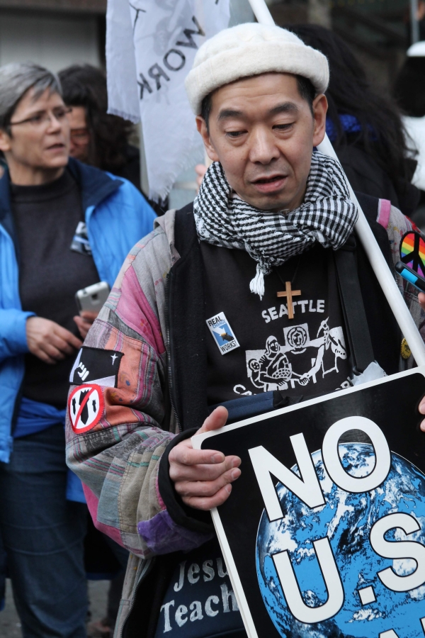 Middle-aged Asian man in cap, scarf, and barn jacket holds sign that says "No U.S. War."