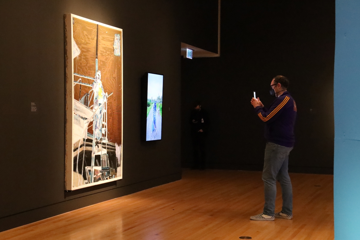 A man stands in a darkened room in front of a painting, holding up his phone.