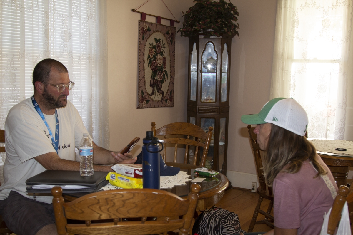 A white man in glasses sits at a dining-room table, which holds papers and water bottles, across from a white woman in a baseball cap.