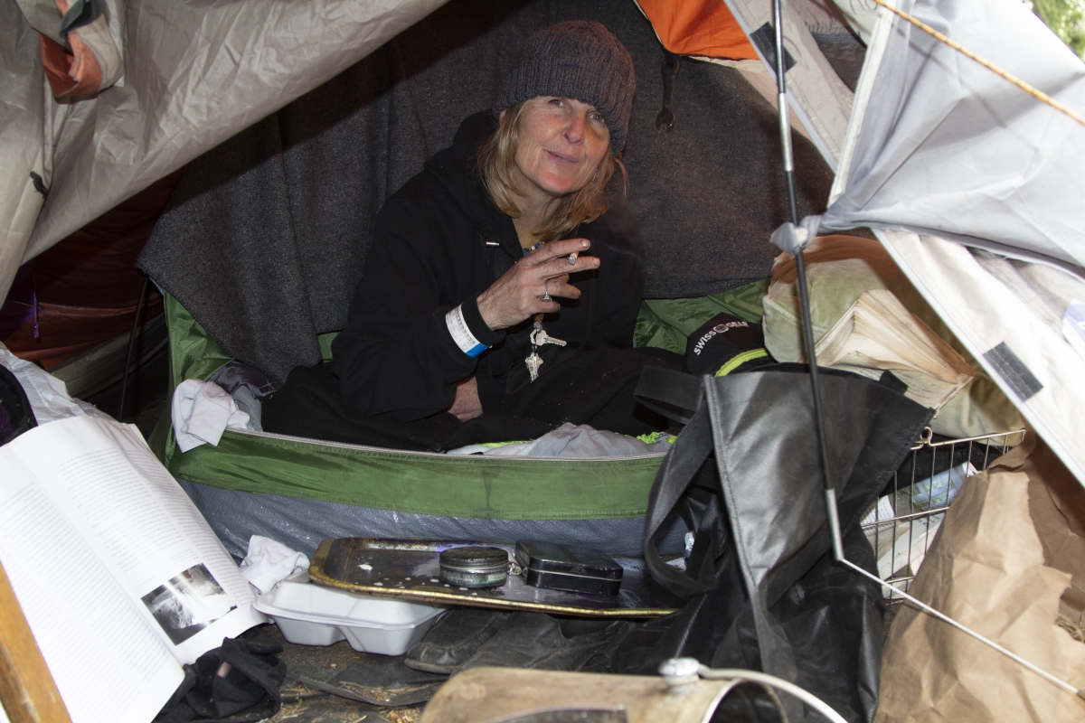 Middle-aged white woman in knit cap and jacket sits in tent holding cigarette and smiling.