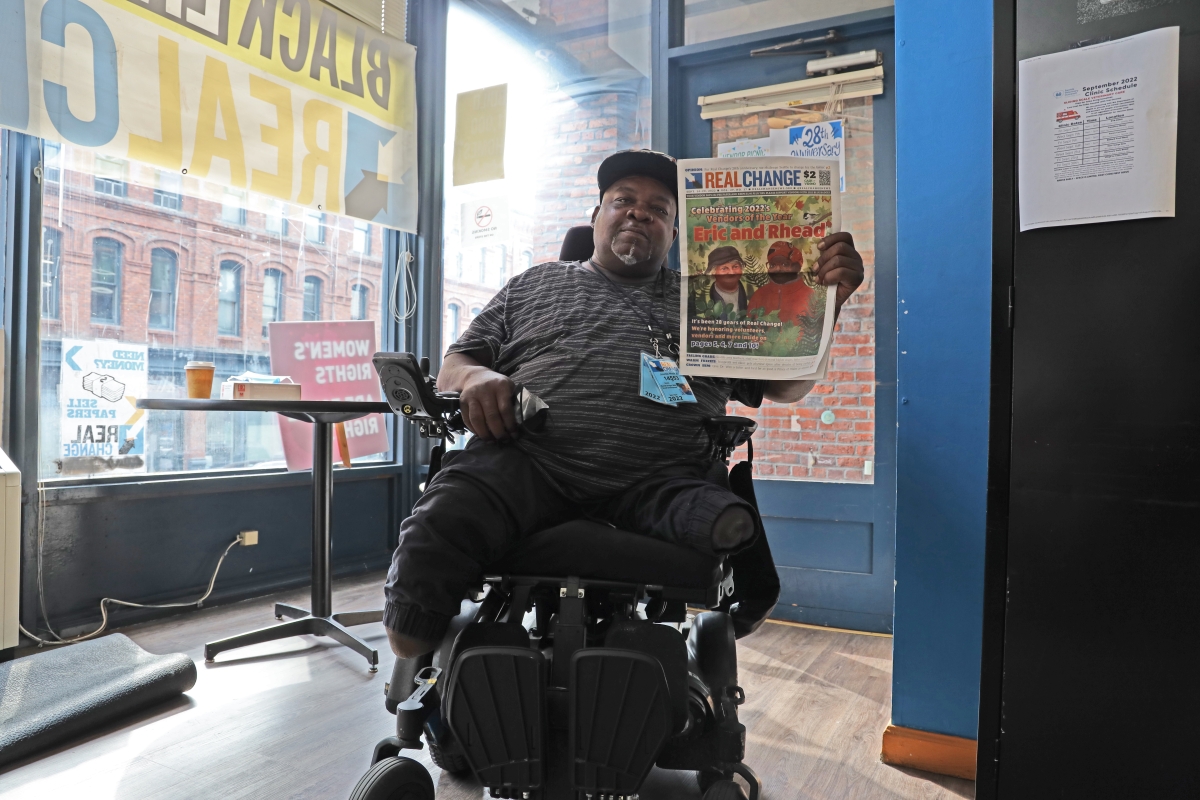 Dark-skinned man in black cap and clothes sits in wheelchair holding up Real Change newspaper.