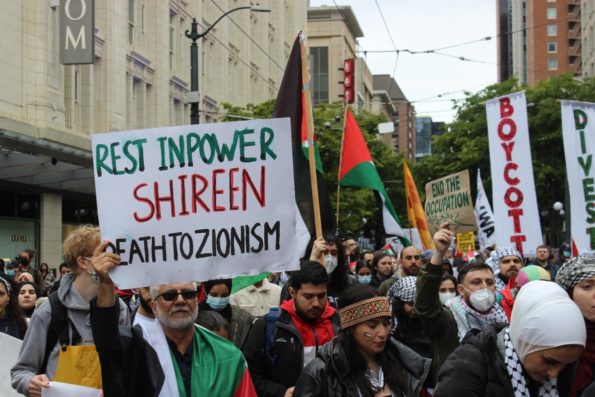 Crowd gathered on city street holding signs; one in foreground reads, "Rest in Power Shireen Death to Zionism"