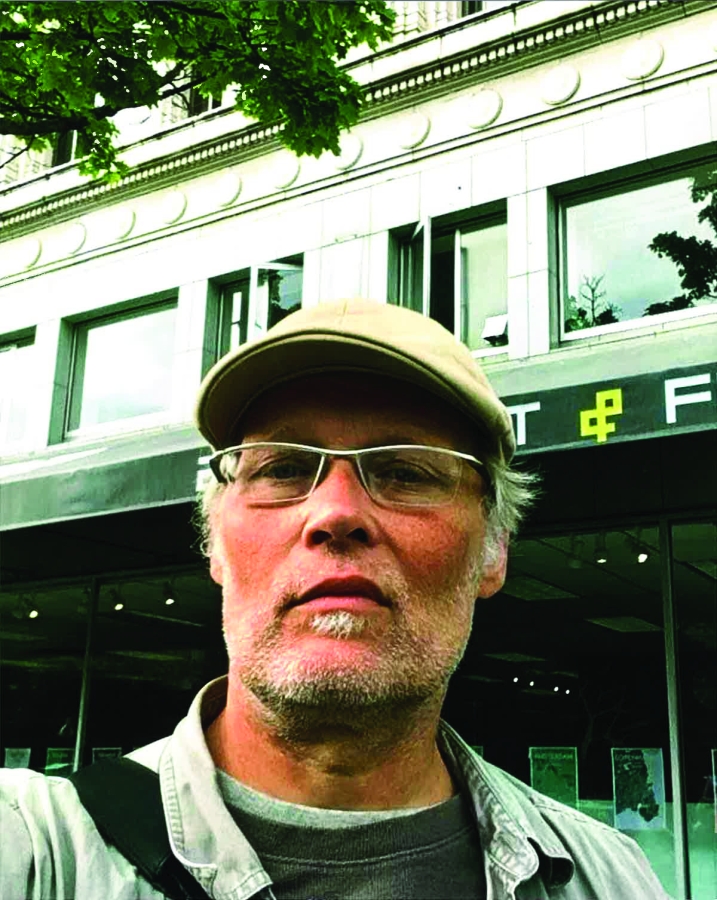 White man in glasses, wearing white goatee, beard, and cap stands outside a building