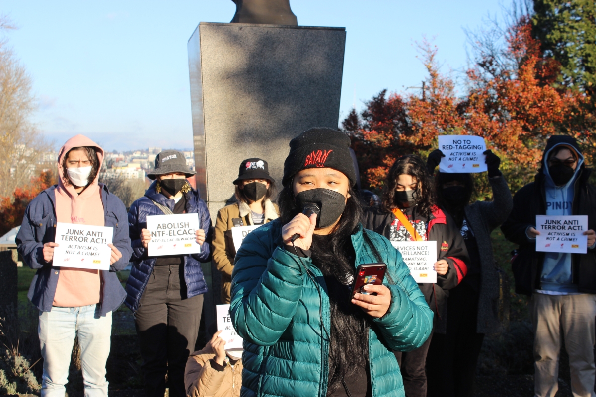 Several young Filipino people, in hats, coats, and masks, stand by a stone monument holding signs on a sunny day. Young woman with microphone stands in foreground.