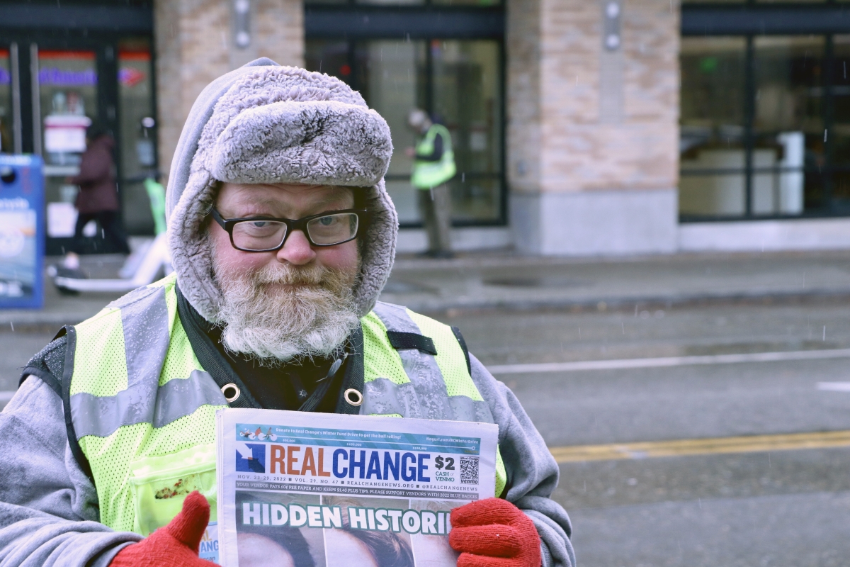 Older white man with glasses and white beard, dressed in ear-flap cap, holds Real Change newspaper wearing sweet expression