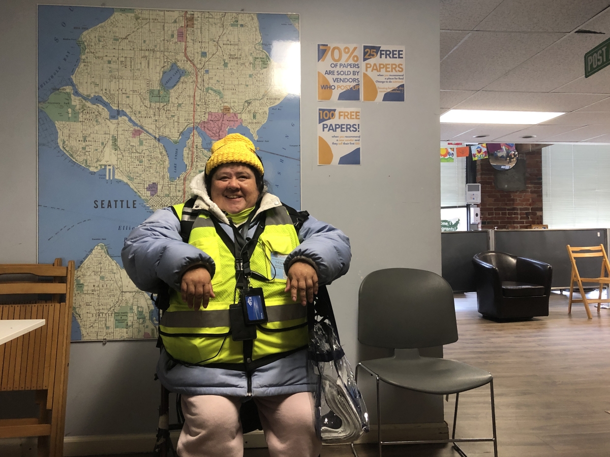 Smiling woman wearing puffy jacket, knit hat, and vendor vest sits in chair in office setting