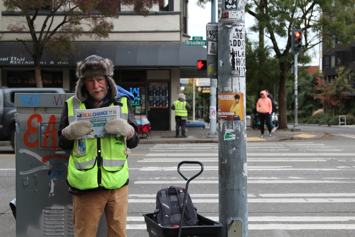Older white man in fur hat with earflaps and vendor vest stands on street corner smiling sweetly while holding up Real Change newspaper