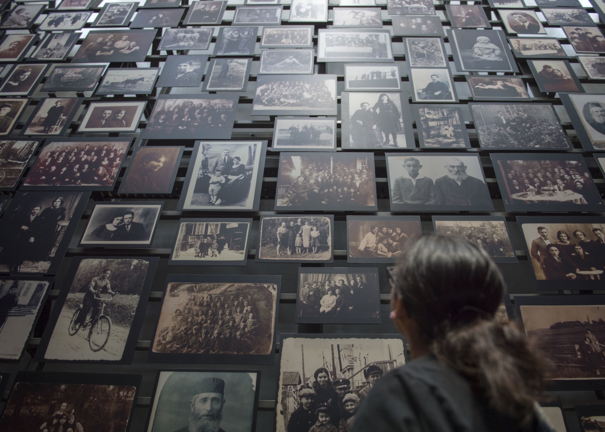 Young white woman wearing glasses and hair in a ponytail, seen from behind, gazing up at a wall of family photos from 1930s and before
