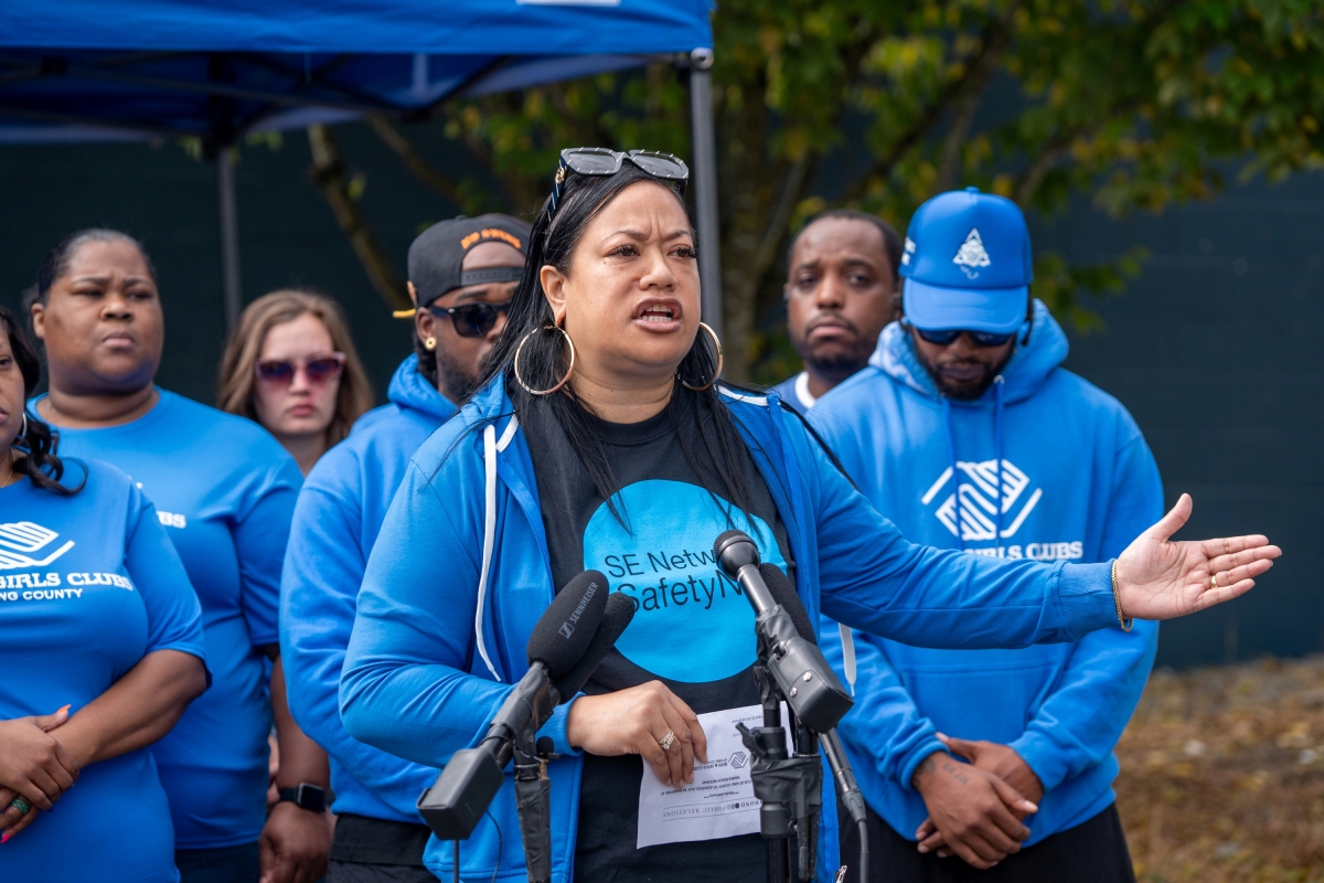 Middle-aged Latina woman gestures and speaks into microphones in front of group of younger people, everyone dressed in matching blue hoodies. 
