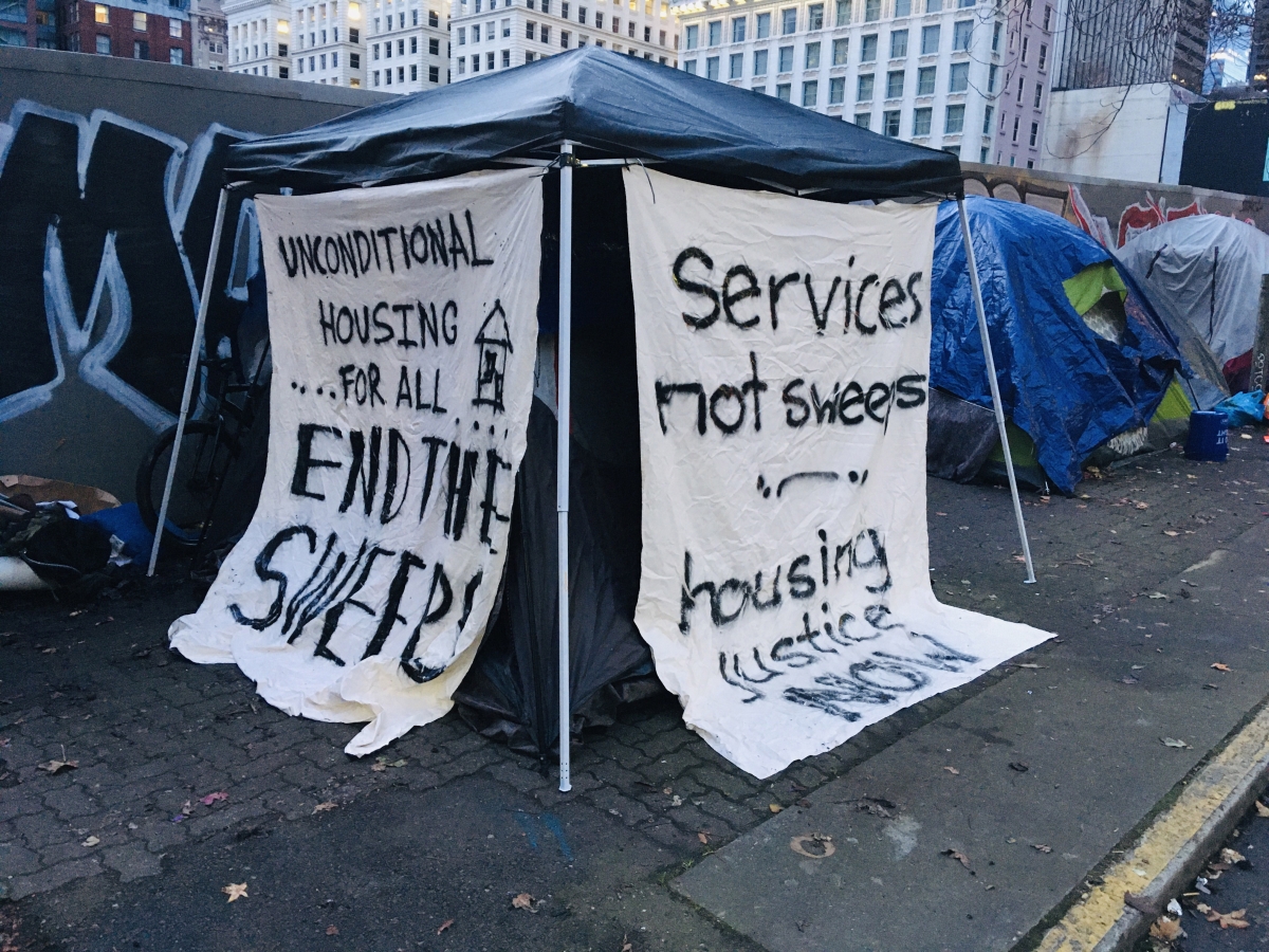 Photograph of tents along city sidewalk festooned with anti-sweeps signs