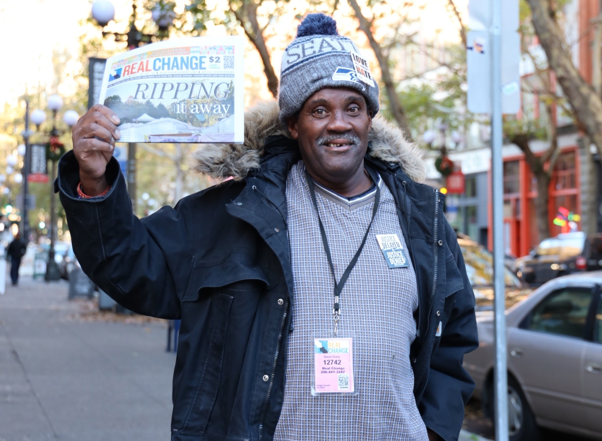 A Real Change vendor holds up the paper, smiling.