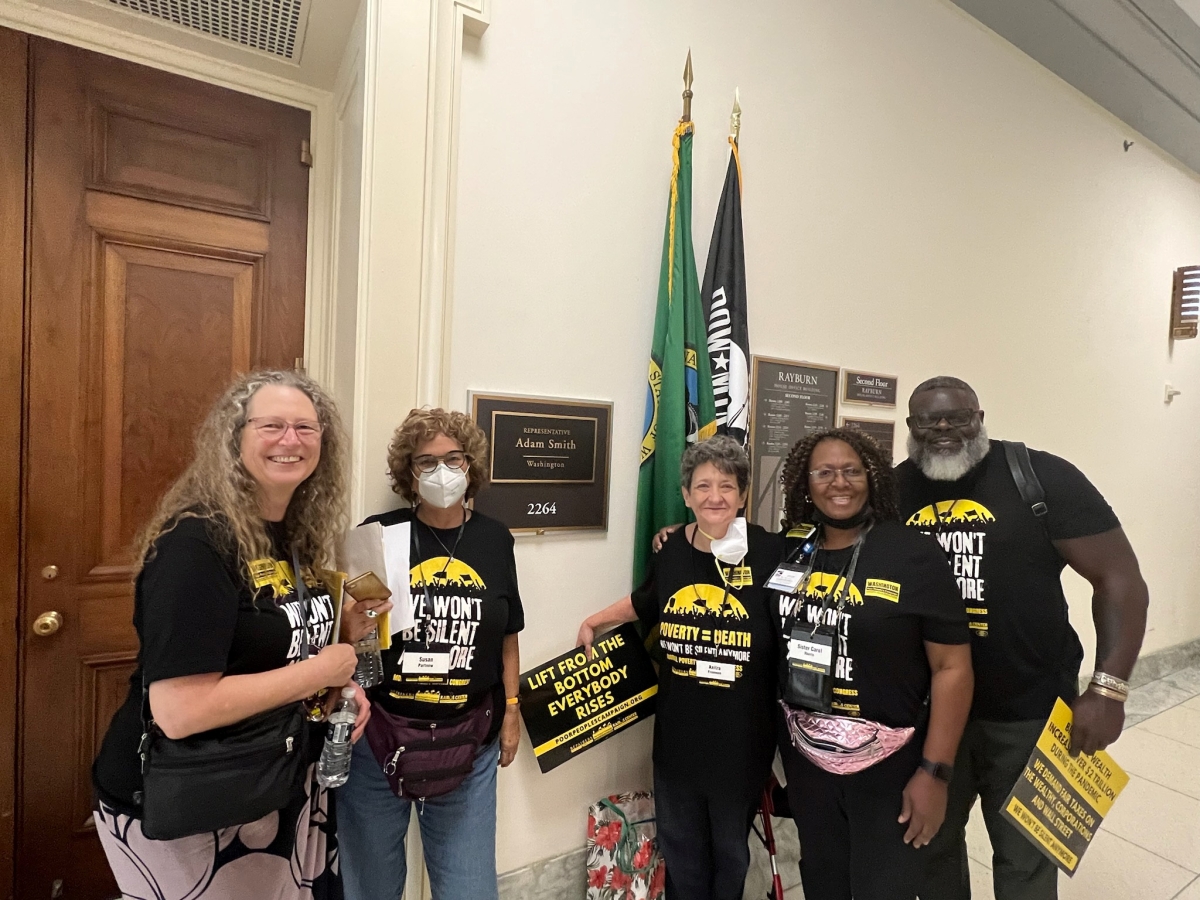 Three middle-aged white women, a Black woman, and a Black man wearing T-shirts reading, "We Won't Be Silent" stand outside a U.S. congressperson's door