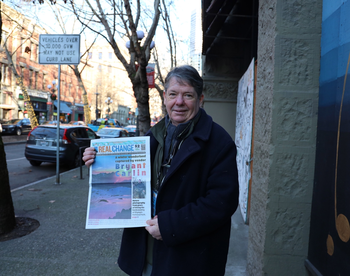 Bryant Carlin, a middle-aged white man wearing a dark overcoat, stands on street corner holding a copy of Real Change featuring his art and name on cover.