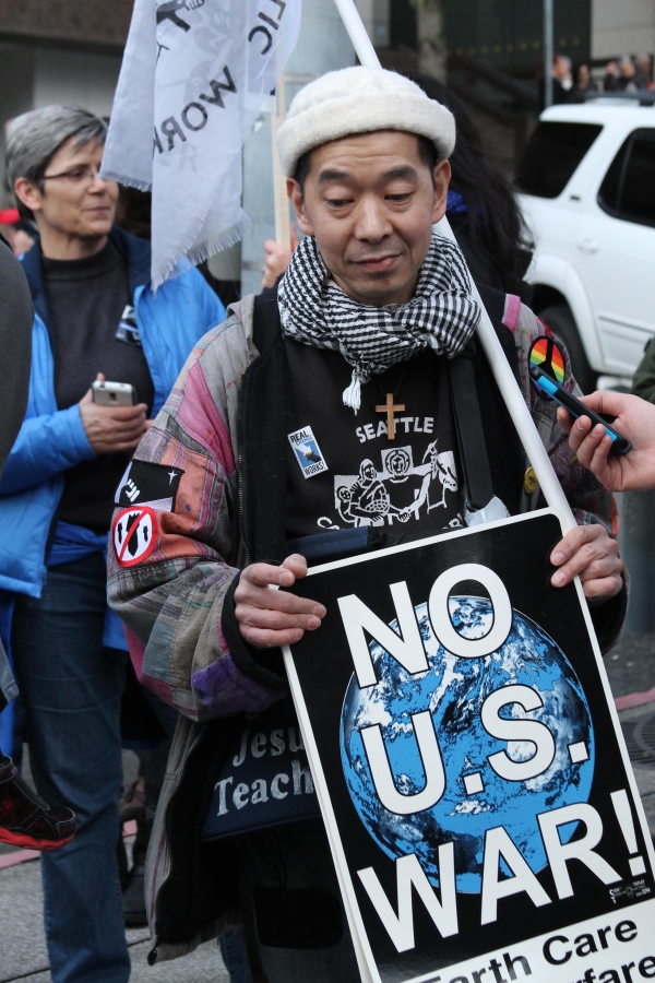 Middle-aged Asian man in cap, scarf, and barn jacket holds sign that says "No U.S. War."
